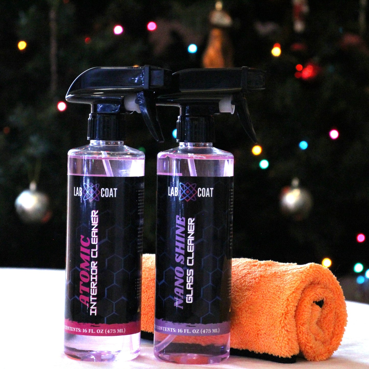 Two spray bottles and an orange towel on a surface with a Christmas tree in the background.