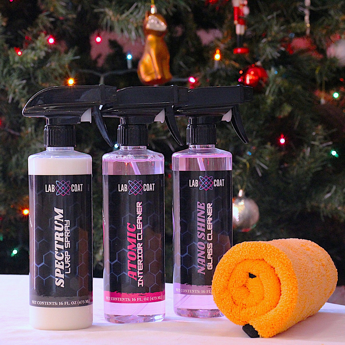 Three bottles of LabCoat cleaning products with a yellow towel on a surface in front of a decorated Christmas tree.