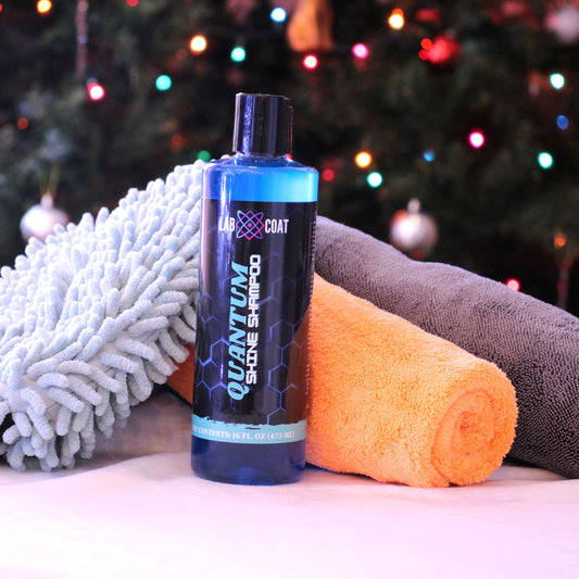 Car care products including a bottle of 'Quantum' car shampoo, microfiber cloths, and a foam brush on a surface with a Christmas tree in the background.
